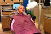 A woman sitting in a dentist s chair, wearing a purple dental hygiene cover and using an oral suction device.