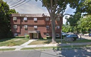 This image is a photograph of a red brick building with multiple windows and two visible entrances, situated in an urban residential area with trees and grass.