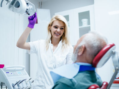 A dental hygienist is holding a mirror over an elderly man's face, both in medical gowns with masks and gloves.